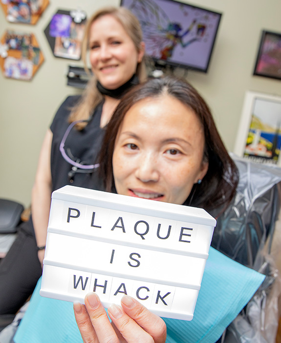 Example of professional photography done for OKC business Masterpiece Smiles. A female patient holds up a sign that reads 'plaque is wack' while a dental hygienist behind her smiles.